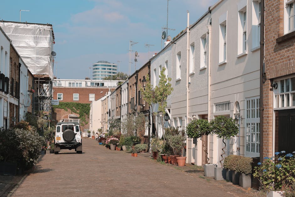 An empty residential street in Kennington with a row of Victorian-style terraced houses featuring bay windows, decorative facades, and brick chimneys, under a cloudy sky. The pavement runs along the r