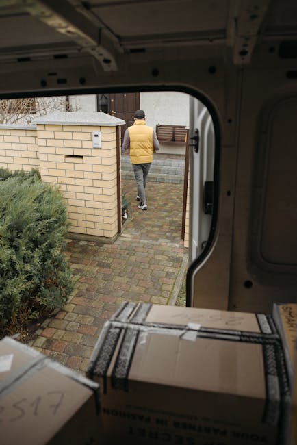 A person’s hand is shown writing the word 'KITCHEN' on a large cardboard box with a black marker, likely as part of packing for a home relocation or moving process. Surrounding the main focus are addi
